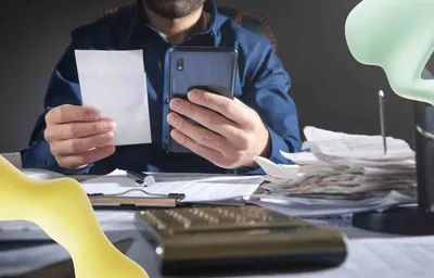 Person scanning a receipt with a smartphone at a messy desk full of papers and a calculator, partially obscured by abstract forms.