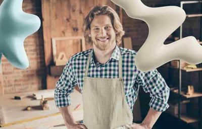 A happy male artisan in a work apron smiles in his studio, with light blue and cream abstract shapes floating around him.