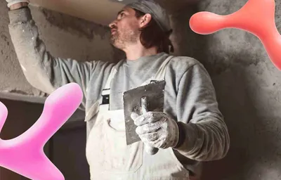 Man in work clothes and gloves applying plaster with a trowel to a wall, partially obscured by large pink and red abstract shapes.