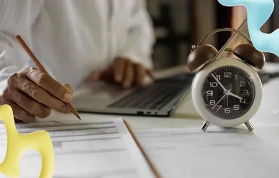 A person's hand writes with a pencil on documents. A laptop is in the blurred background, with a white alarm clock on the desk.