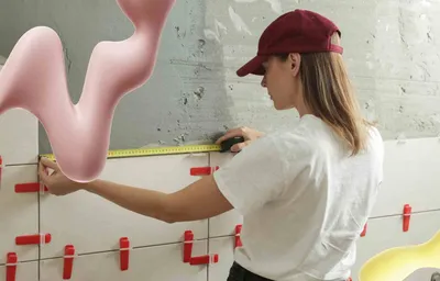 A woman in a red cap measures white wall tiles with a tape measure. Pink and yellow wavy abstract shapes float in the scene.