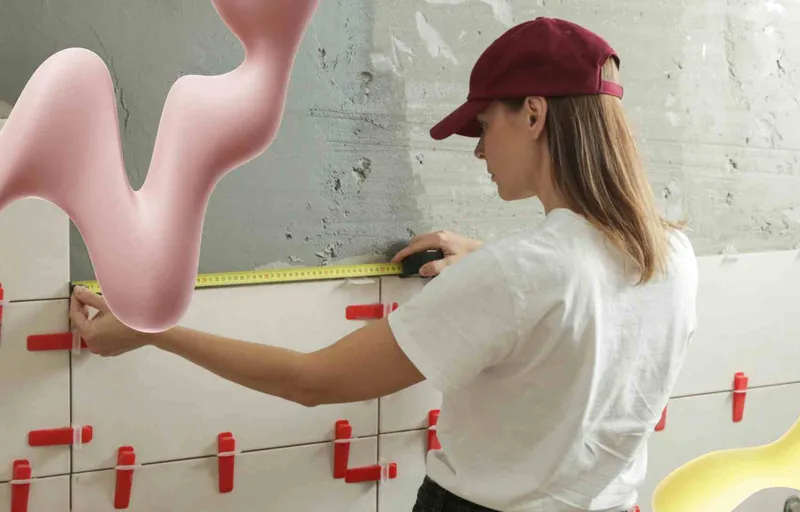 A woman in a red cap measures white wall tiles with a tape measure. Pink and yellow wavy abstract shapes float in the scene.