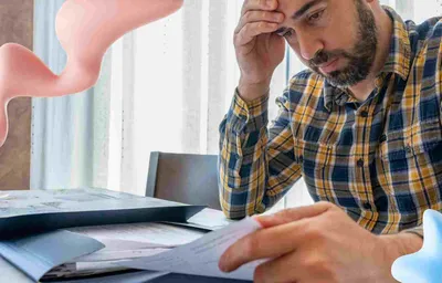 Stressed bearded man in a plaid shirt reading documents at a table, with abstract pink and blue shapes.