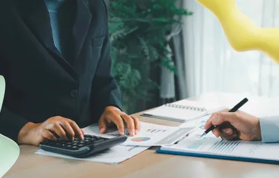Two individuals at a desk, one calculating with a calculator, the other writing on business charts. Financial analysis.