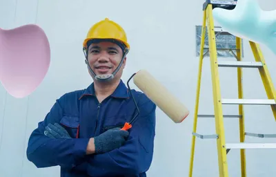 A smiling man in a yellow hard hat and blue uniform holds a paint roller, with a yellow ladder in the background.