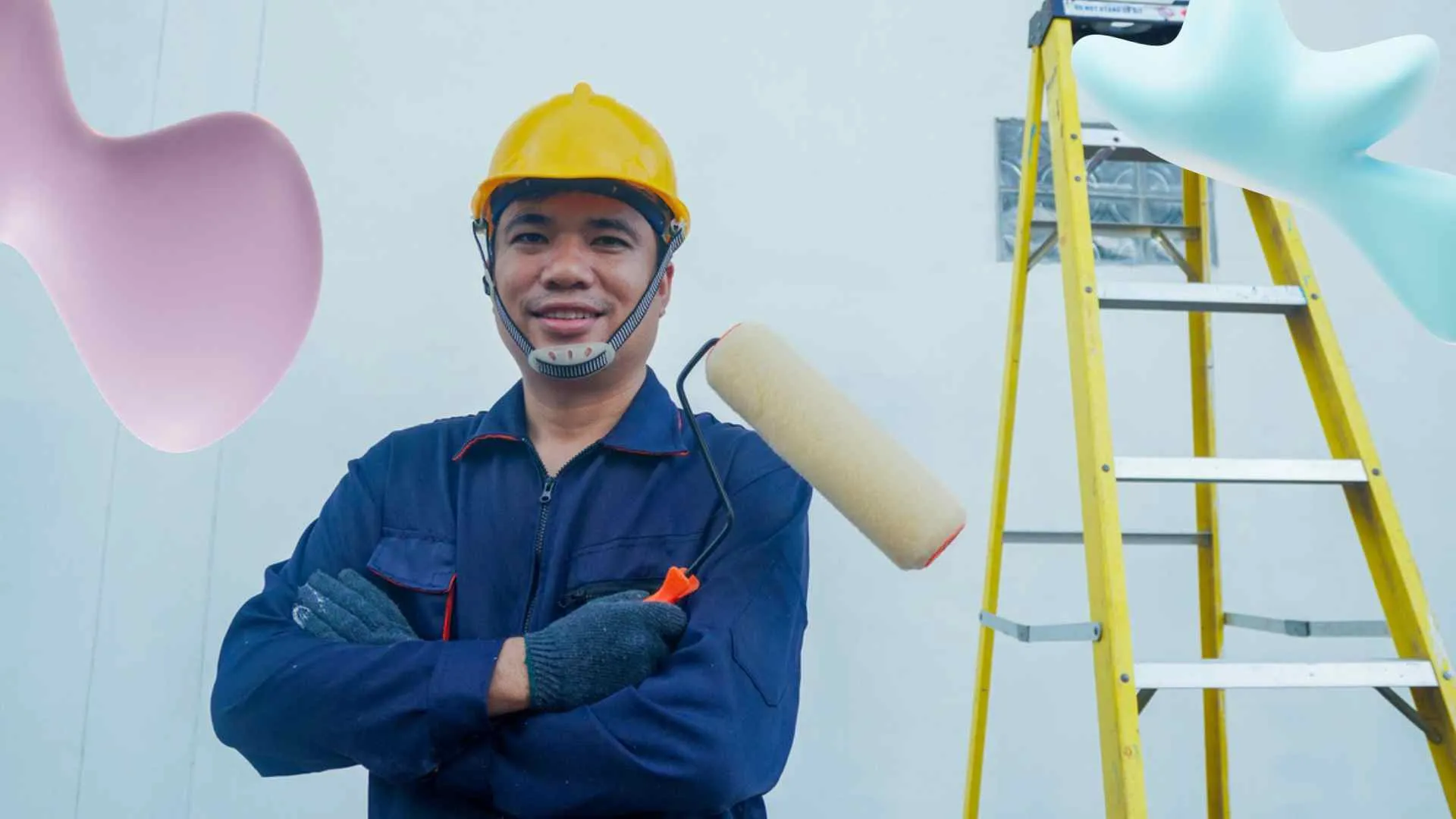 A smiling man in a yellow hard hat and blue uniform holds a paint roller, with a yellow ladder in the background.