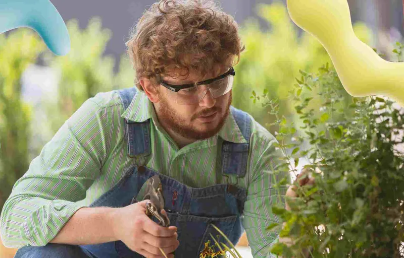 A man in denim overalls, green shirt, and safety glasses prunes a green plant with shears, focused on his task.