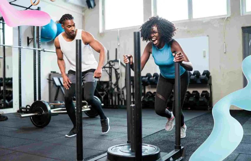 A woman with an intense, yelling expression pushes a heavy sled in a gym. A man walks behind her, also exercising.