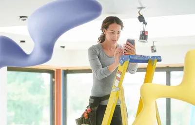 A woman on a ladder with a tool belt uses her smartphone while working on ceiling lights, flanked by abstract purple and yellow shapes.