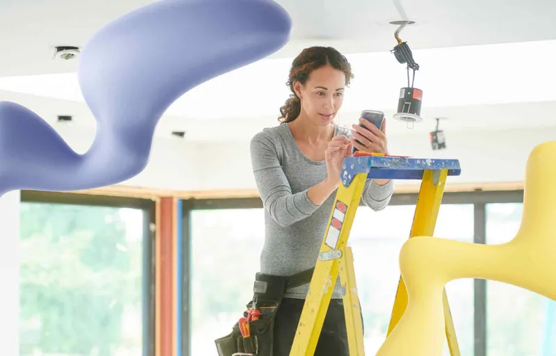 A woman on a ladder with a tool belt uses her smartphone while working on ceiling lights, flanked by abstract purple and yellow shapes.