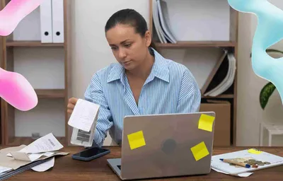 Woman examining receipts at a cluttered desk with a laptop, paper, and phone. Abstract pink and blue forms hover nearby.