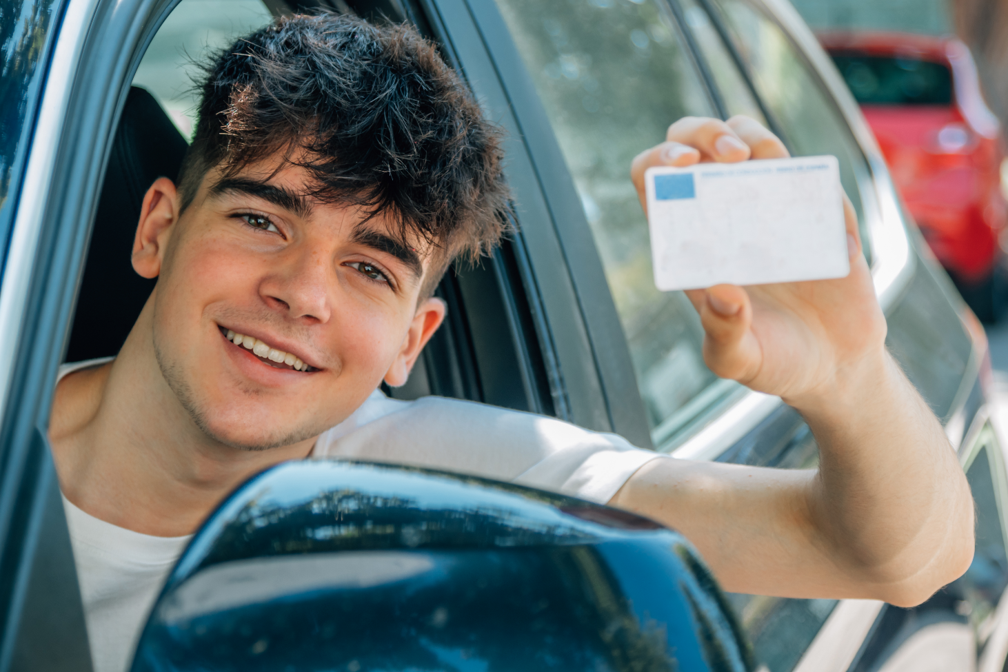 A young man happily looking out of a vehicle's window while proudly showing off his UK driving licence (blurred)