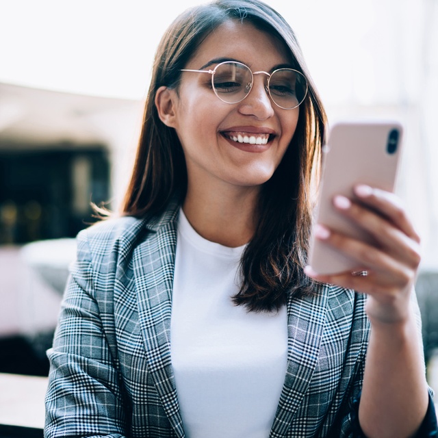LINK Mobility -  Fröhliche junge Dame mit mit Blazer und Brille erhält eine SMS auf ihrem Smartphone