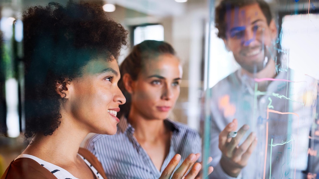 Drei Personen, die in einem Büro Brainstorming betreiben, lächeln. Das Diagramm auf der Glaswand zeigt die bestehenden Opt-in und Opt-out-Prozesse, die nun gemeinsam überprüft werden.