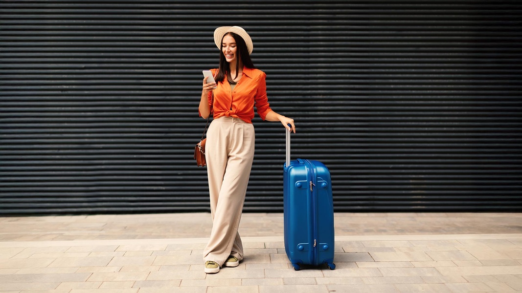 Eine Frau ist auf Urlaub und mit ihrem Koffer unterwegs und lächelt in ihr Handy. Sie erhält eine RCS-Nachricht von dem Hotel, dass sie gebucht hat mit einem Sonderangebot für ein Zimmer-Upgrade.