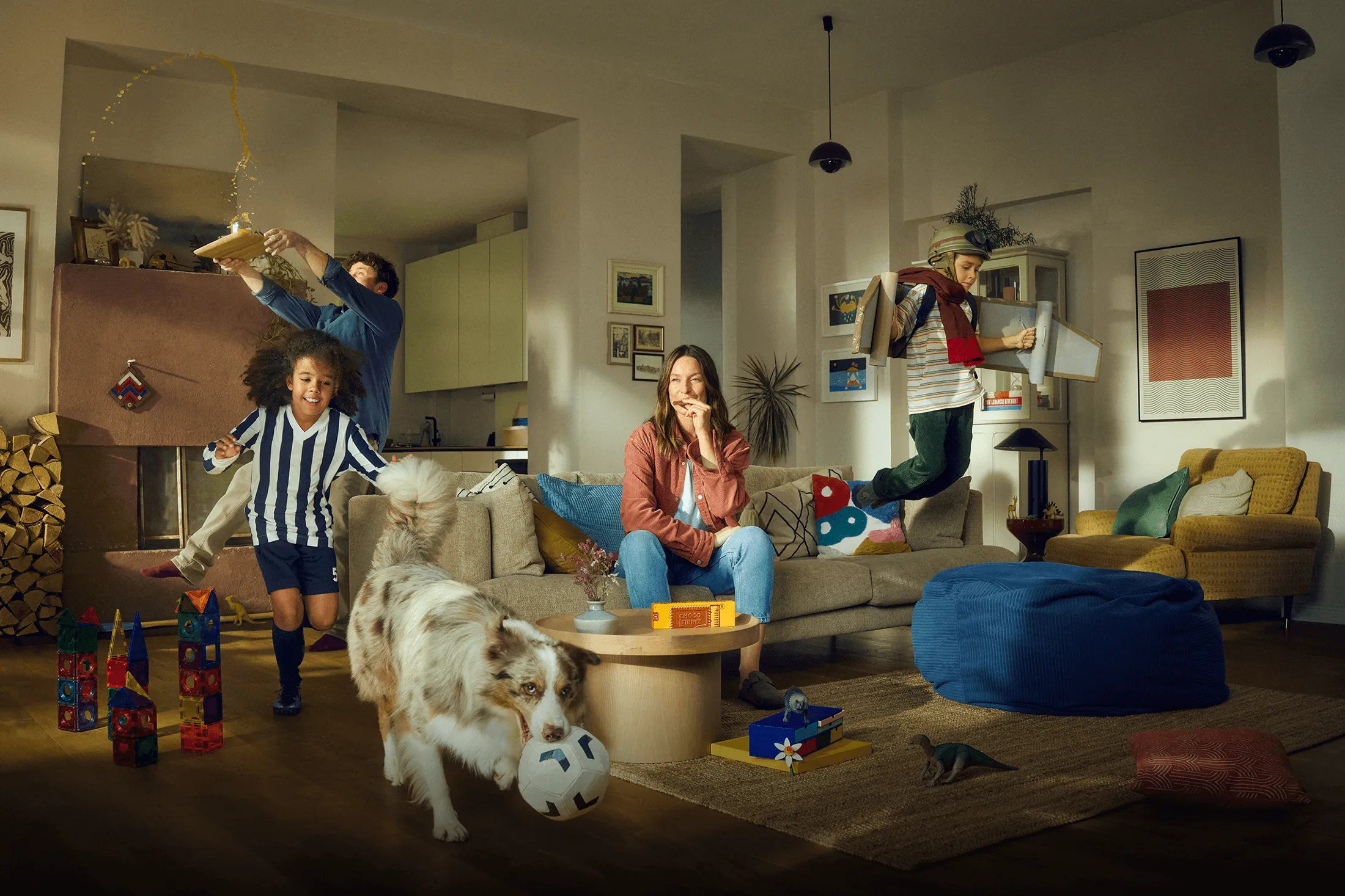 Chaotische Szene im Wohnzimmer einer Familie, während die Mutter auf dem Sofa einen Keks genießt