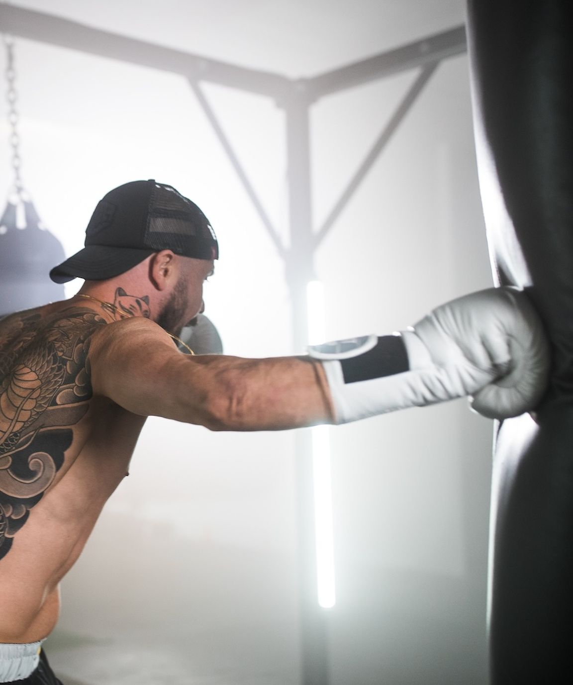 Man with a back tattoo punches a heavy bag in a gym, wearing a cap and boxing gloves.