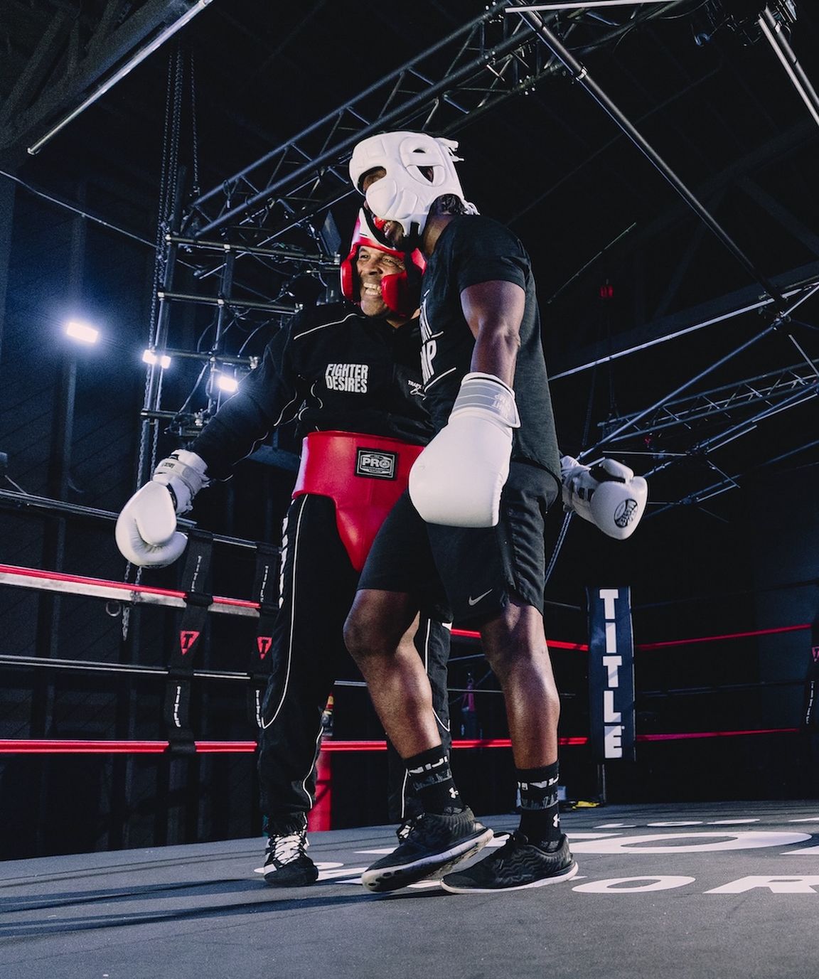 Two boxers in headgear and gloves stand in a ring, under bright lights, with a supportive gesture as they prepare for sparring.