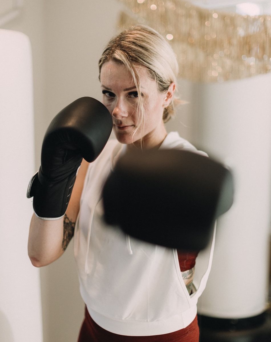 A woman wearing boxing gloves throws a punch, focused and determined, with a blurred background and decorative fringe above.