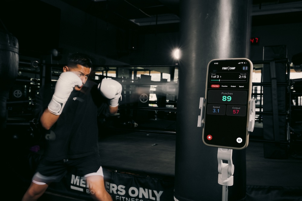 A fighter works the heavy bag in a gym, with the FightCamp app showing his punch stats on a phone