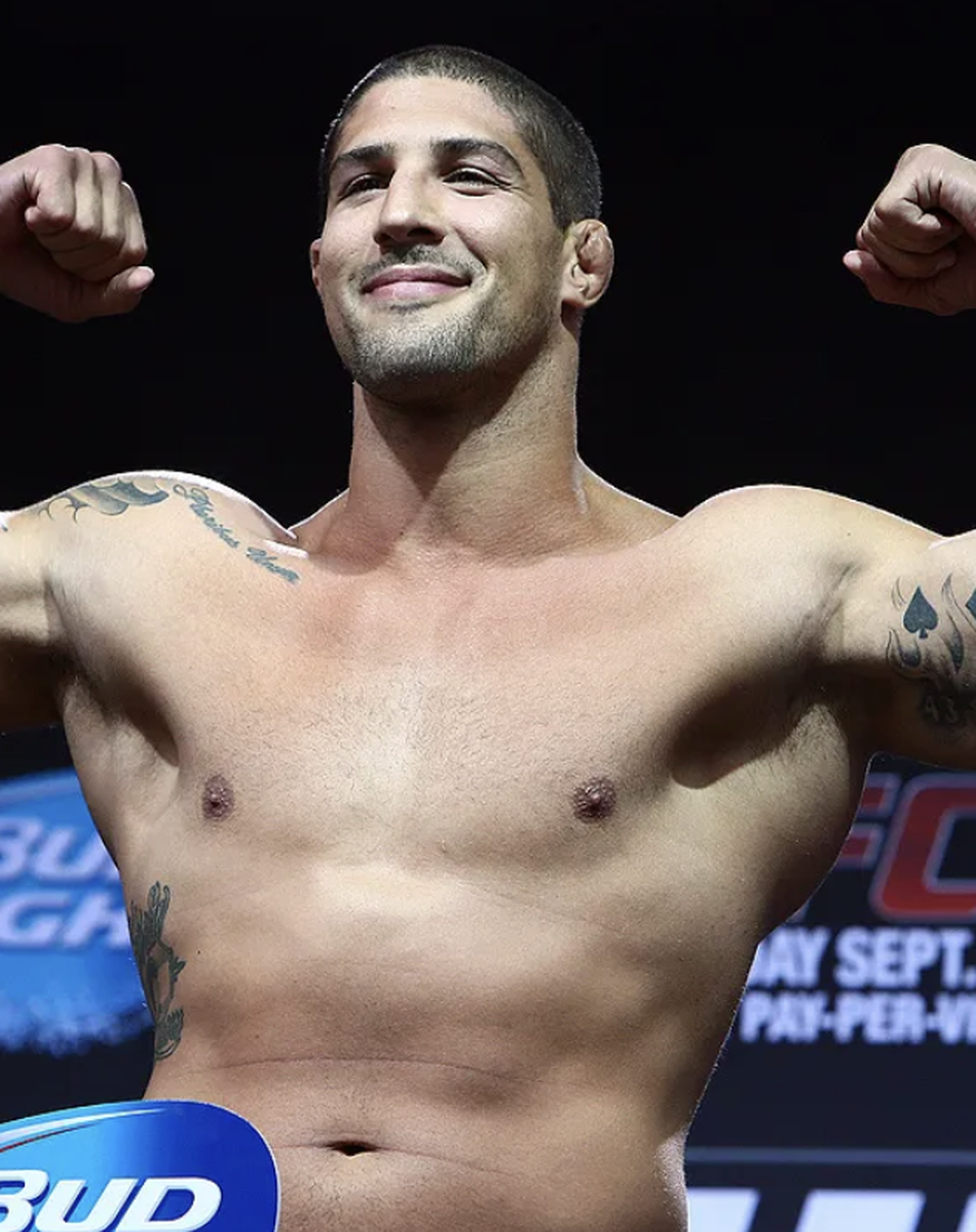 Brendan Schaub flexing his muscles onstage, showing tattoos on arms. He is smiling, and there are advertisements and event date in the background.