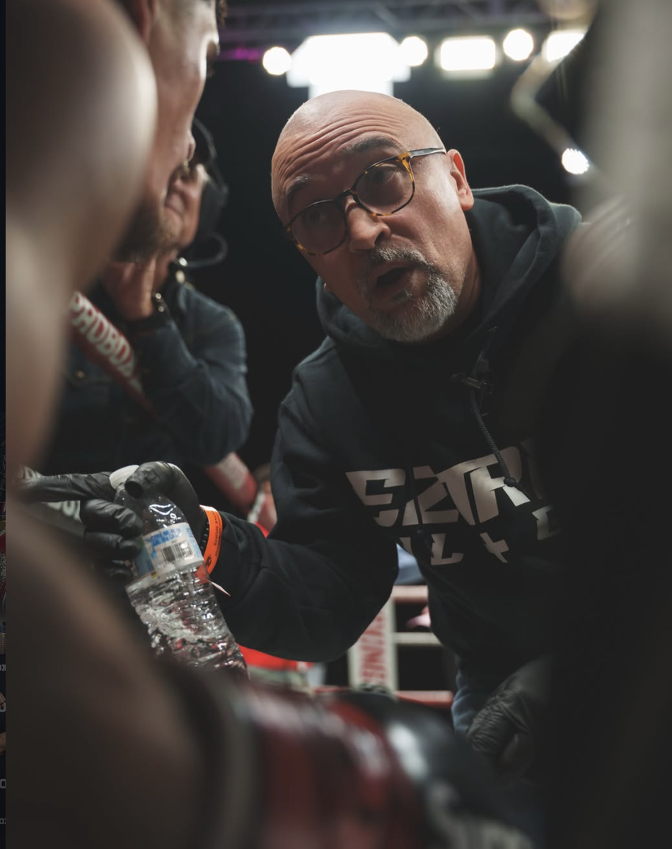 Javiel Centeno wearing glasses and a hoodie, intensely speaks to a boxer, holding a bottle of water, under bright lights in a boxing gym.