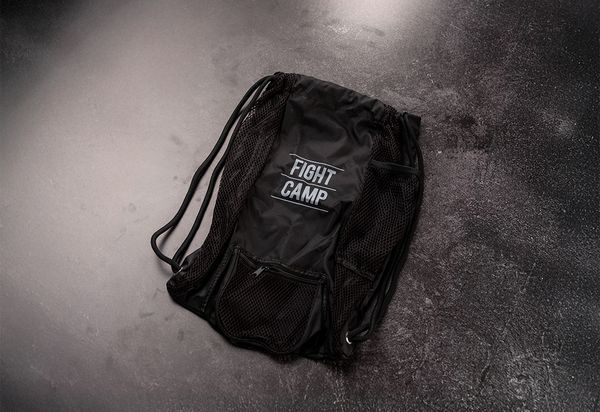 Black drawstring backpack with "Fight Camp" logo on a textured dark surface, featuring mesh pockets and a zippered compartment.
