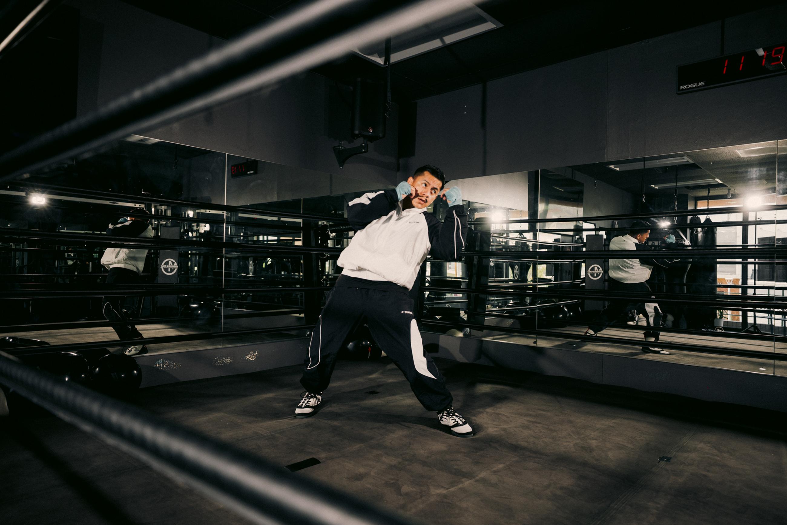 Person in a boxing ring, wearing a tracksuit and hand wraps, practices boxing moves in front of a mirror.