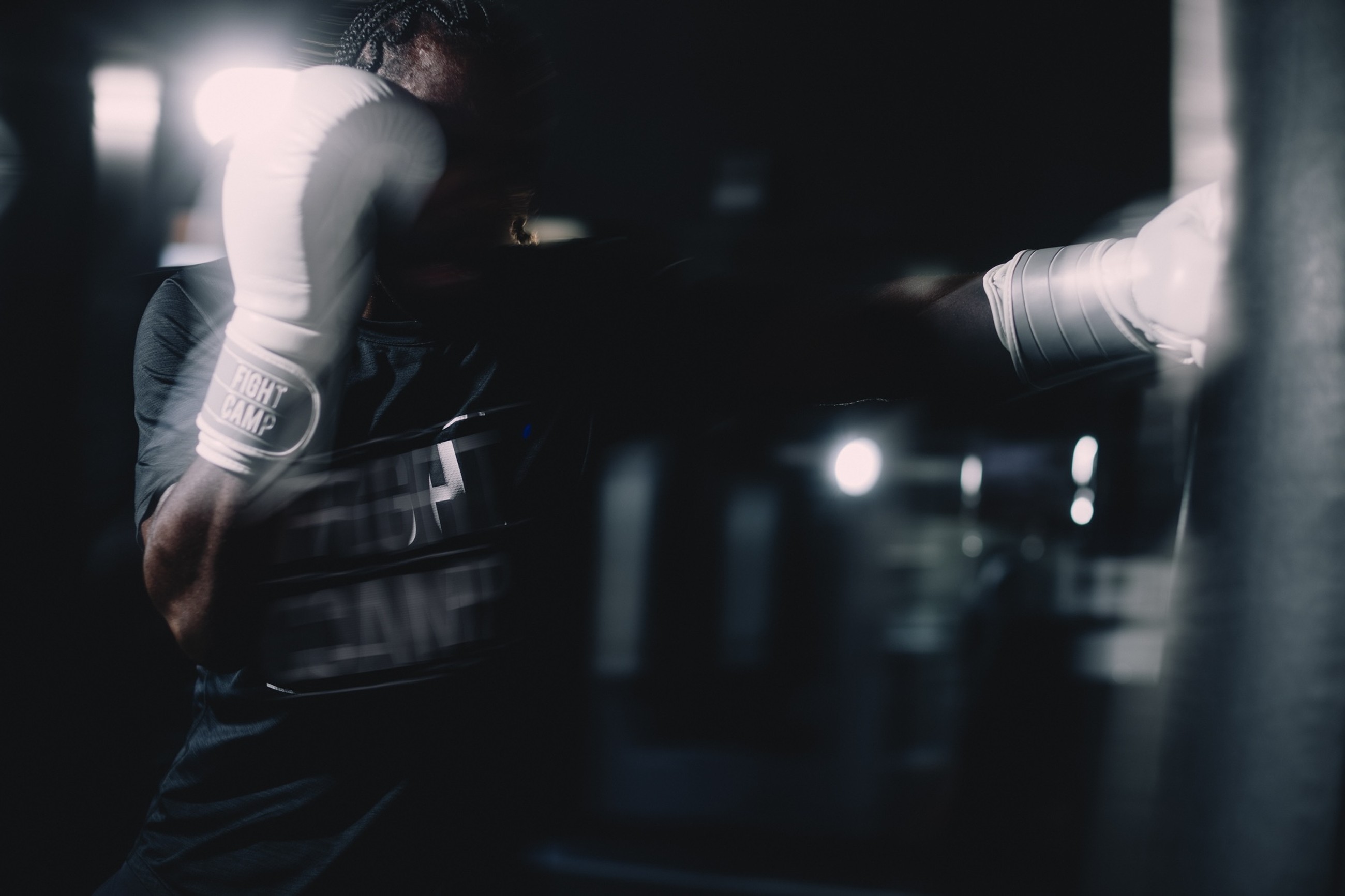 Boxer in dark gym throwing a blurred punch at a punching bag, wearing white gloves and a "Fight Camp" shirt.