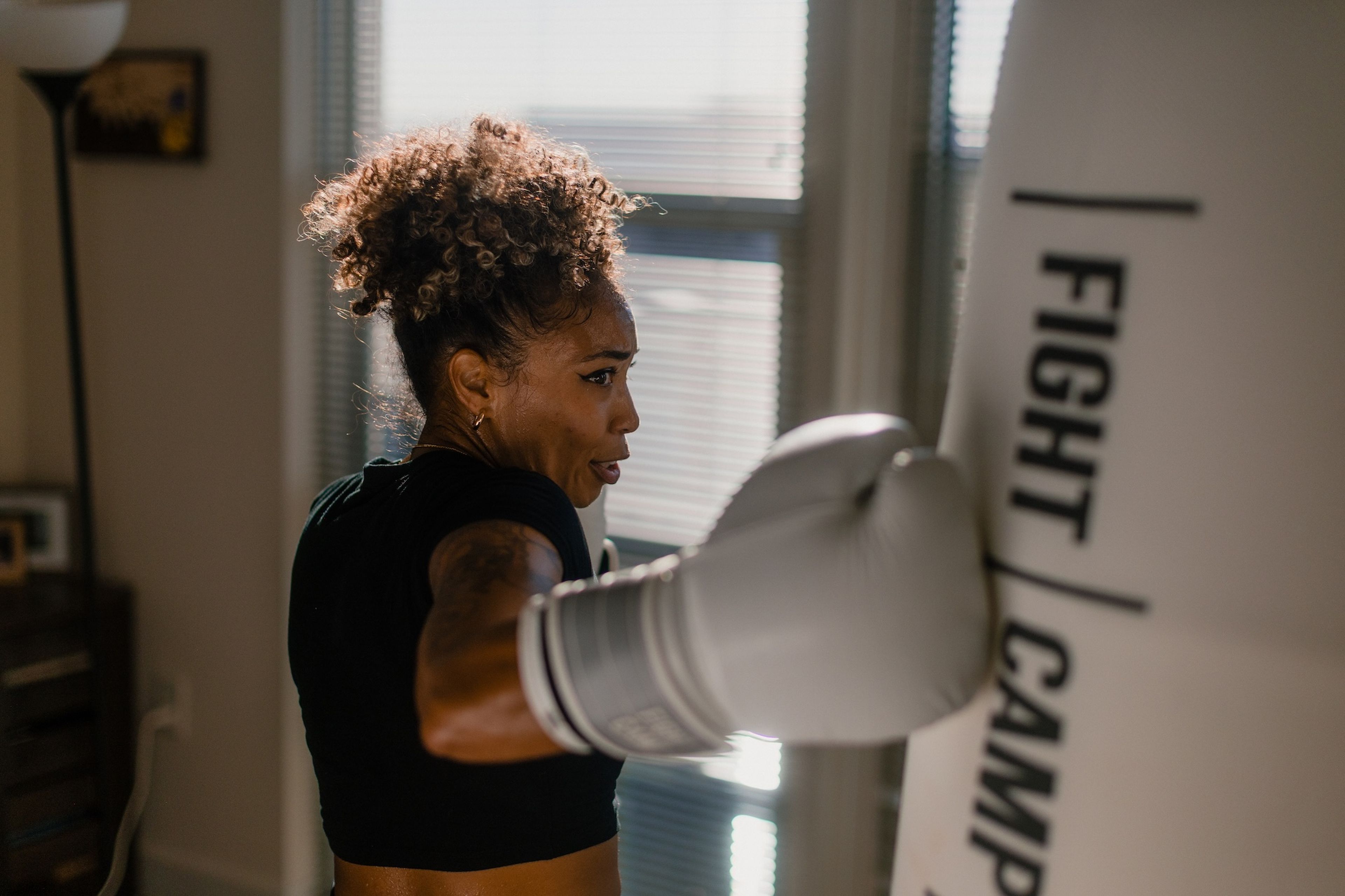 A woman in boxing gloves throws a punch at a punching bag in a sunlit room.