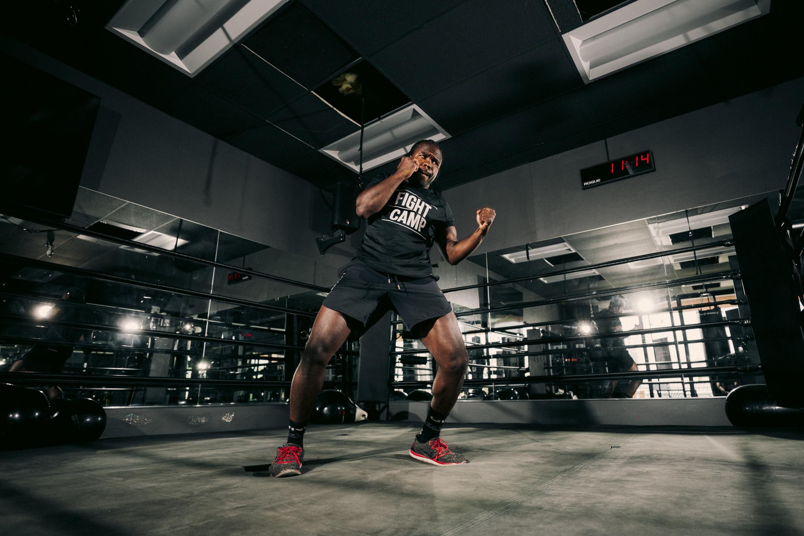 Person in a boxing stance at a gym, wearing a "Fight Camp" shirt, reflected in mirrors.