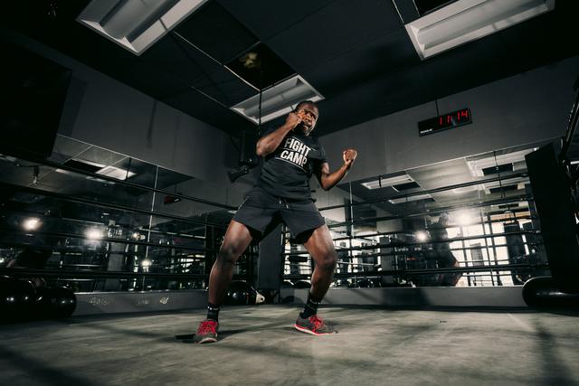Person in a boxing stance at a gym, wearing a "Fight Camp" shirt, reflected in mirrors.