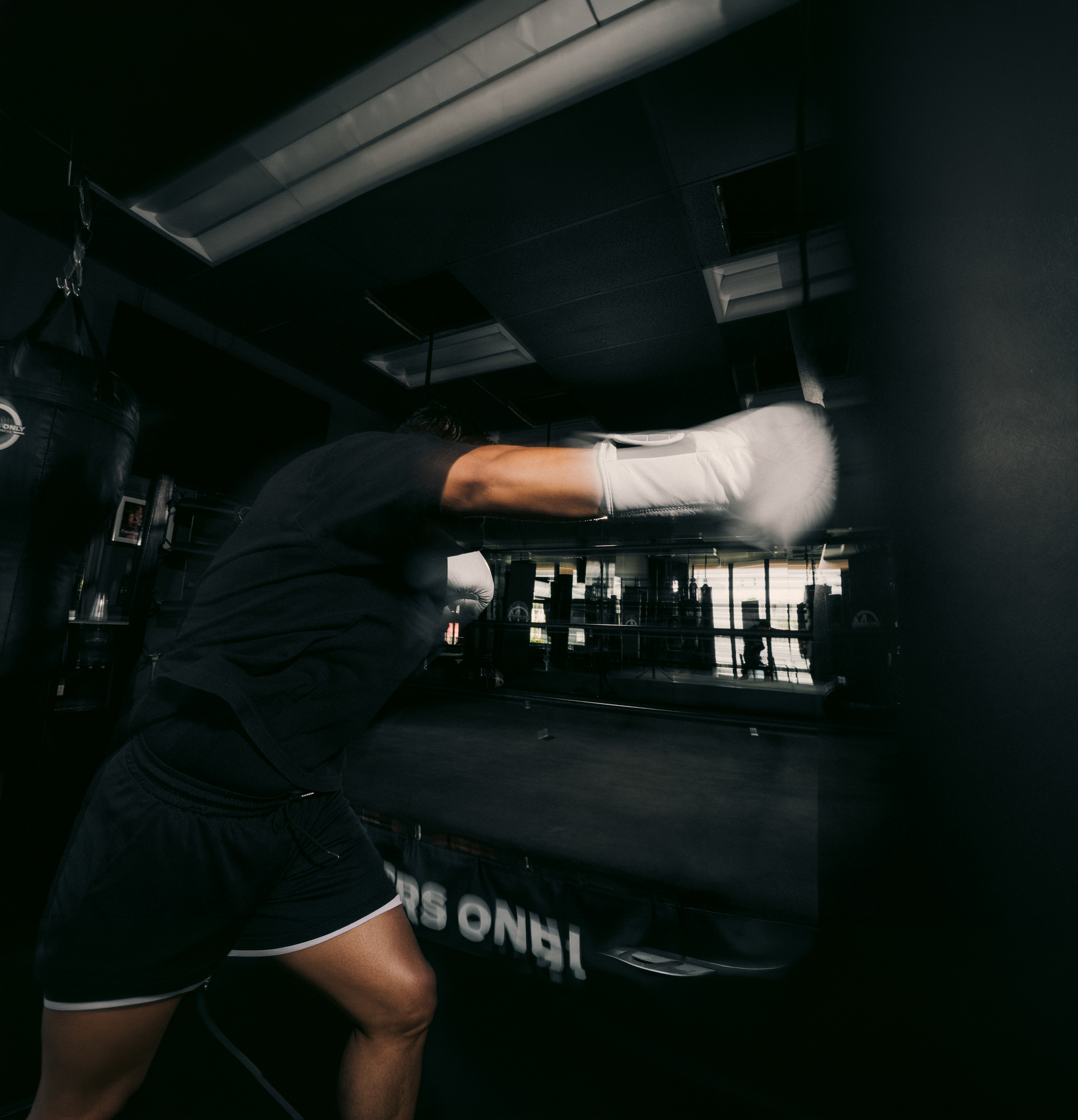 A boxer in black attire throws a punch with white gloves in a dimly lit gym, focusing on a hanging heavy bag.