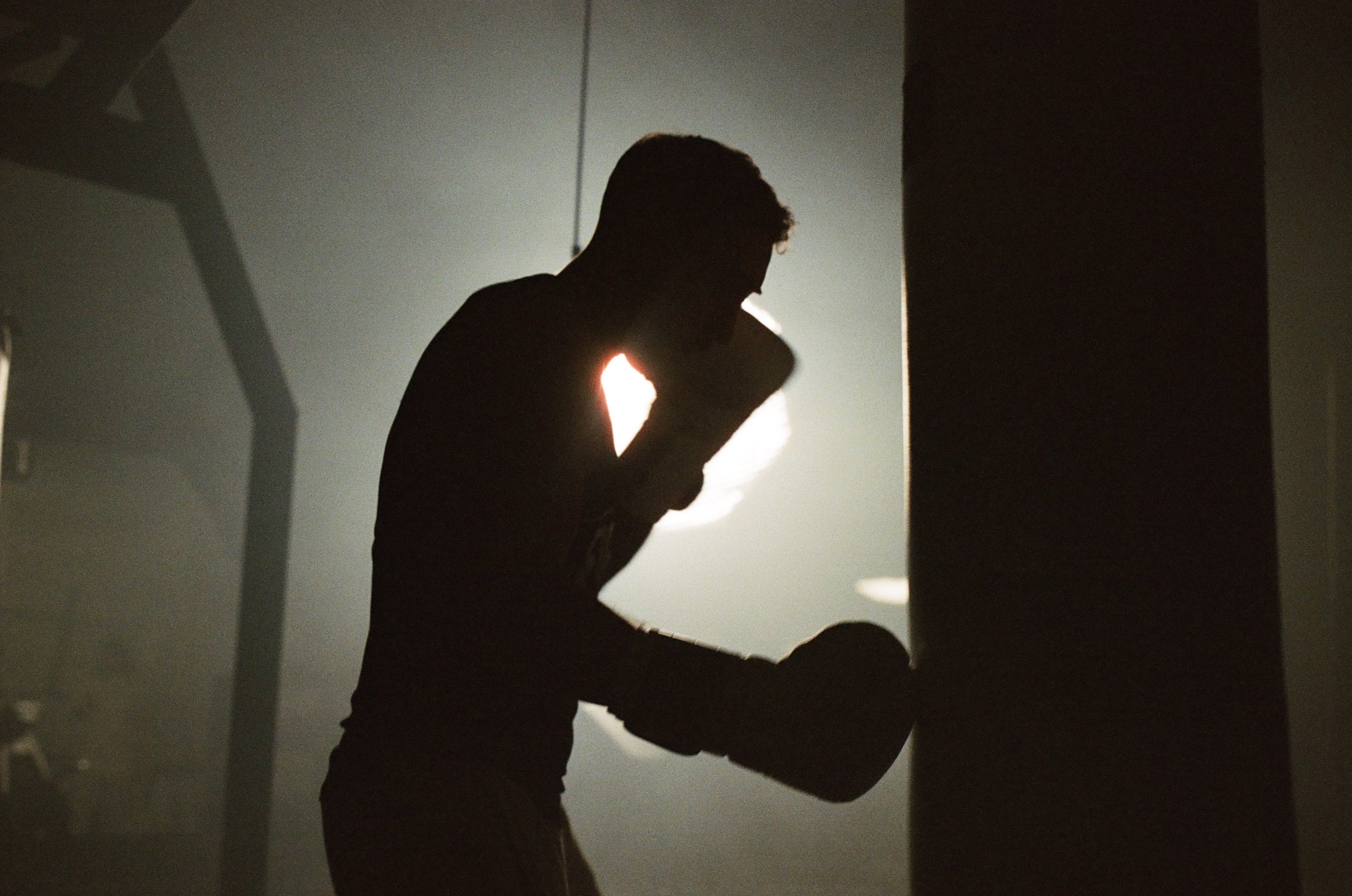 Silhouette of a boxer training with a punching bag in a dimly lit gym, with a bright light in the background.