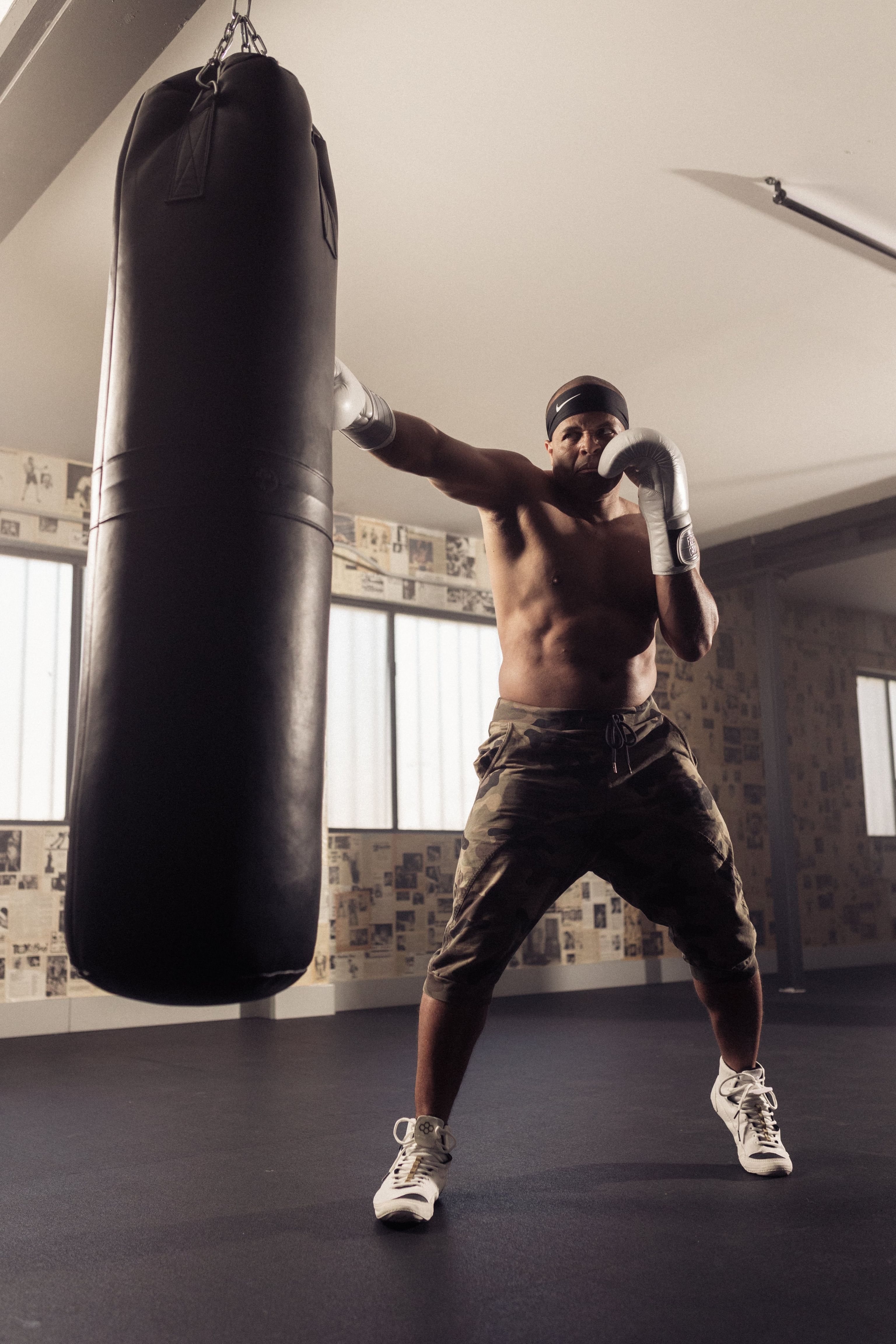 A man in camo shorts and boxing gloves punches a heavy bag in a gym, with large windows in the background.