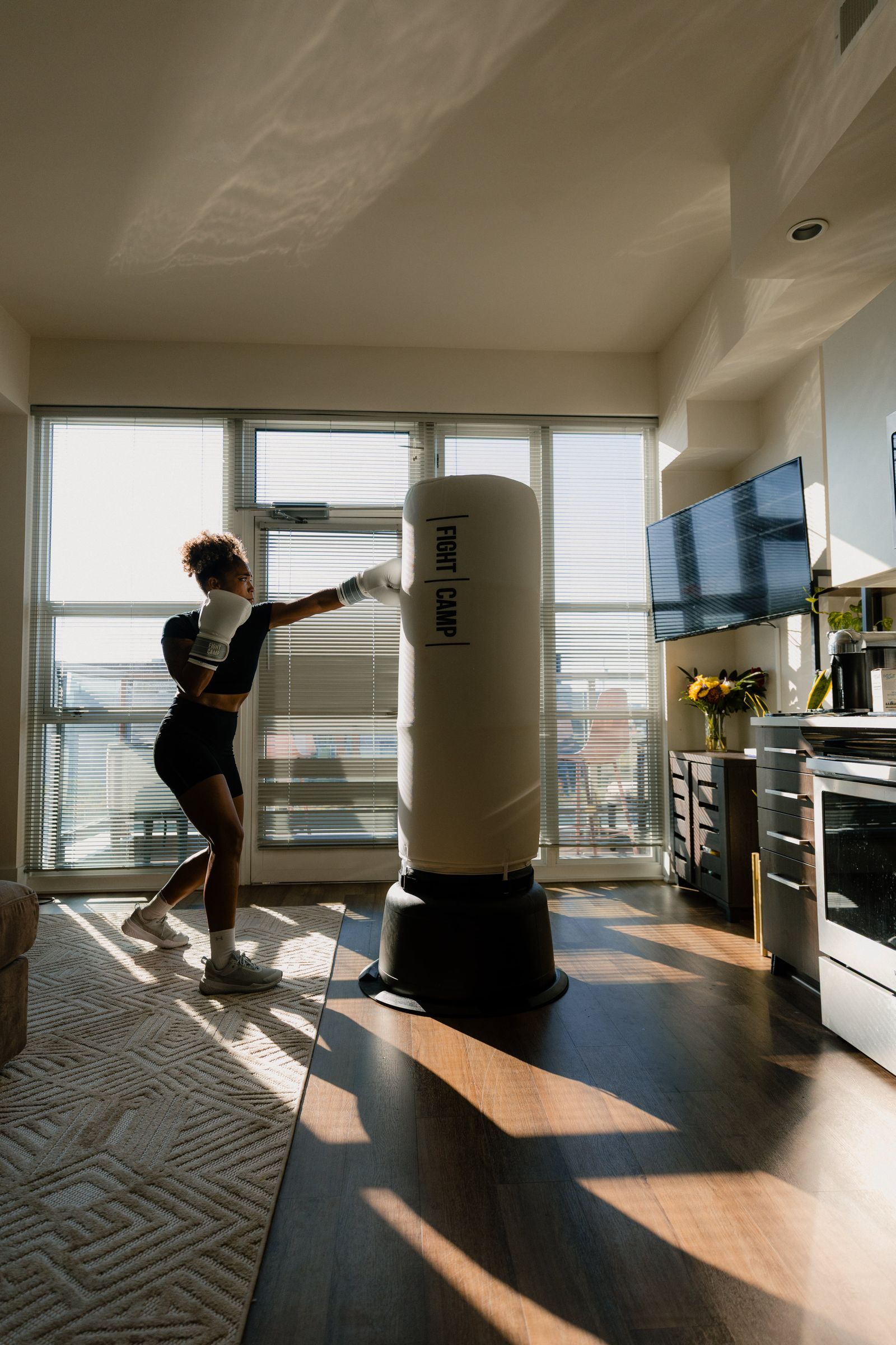 Person boxing a FightCamp punching bag in a sunlit living room, with a TV and kitchen appliances in the background.
