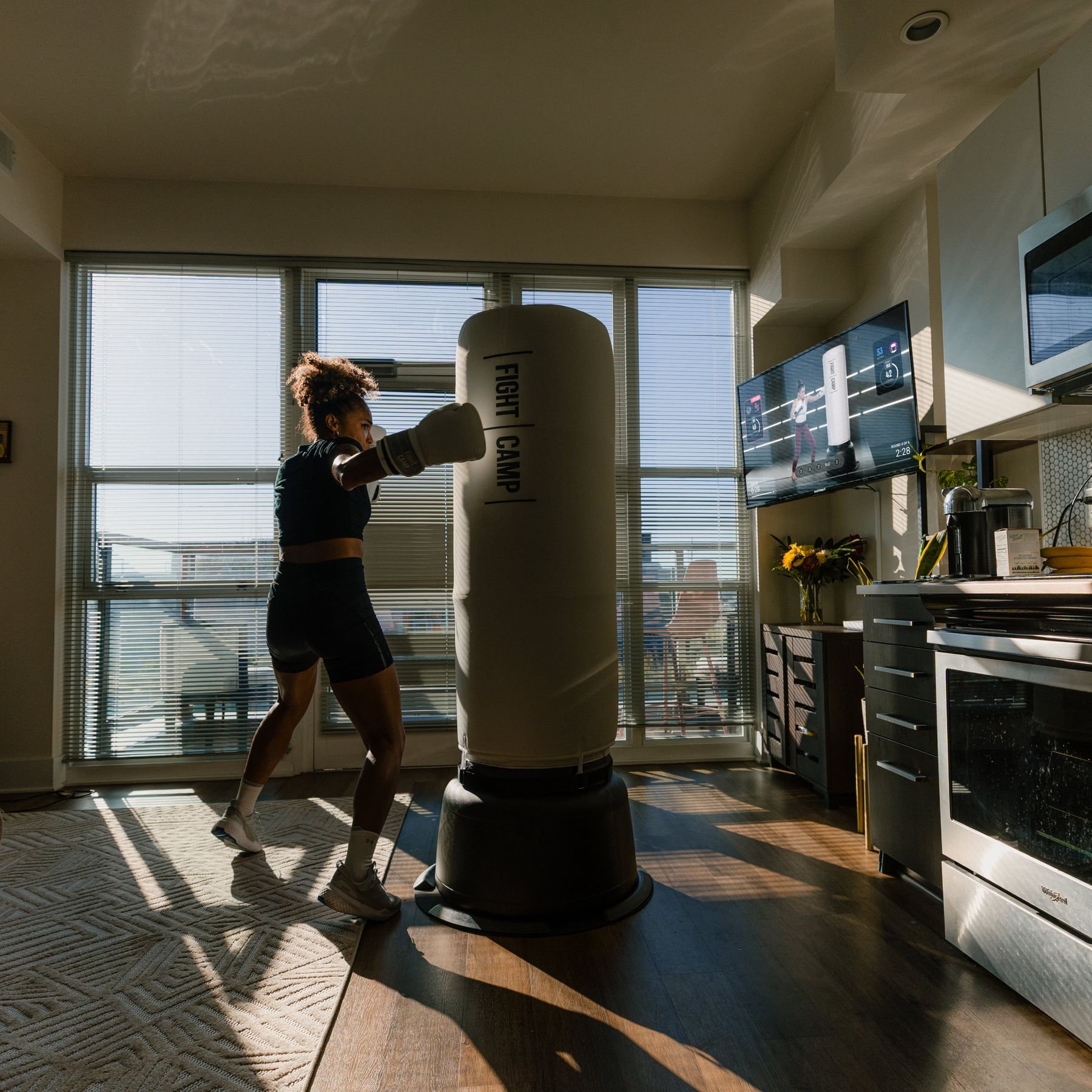 A person in athletic wear practices boxing with a punching bag in a sunlit living room, with a TV displaying a workout video.