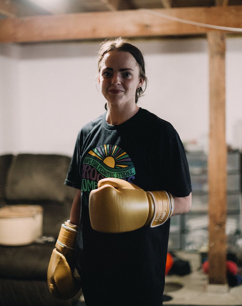 Person wearing boxing gloves and a black T-shirt stands in a room with wooden beams and a couch, smiling at the camera.