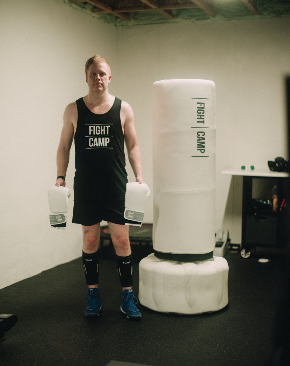 Man in a "Fight Camp" tank top holds boxing gloves, standing next to FightCamp punching bag in a home gym.