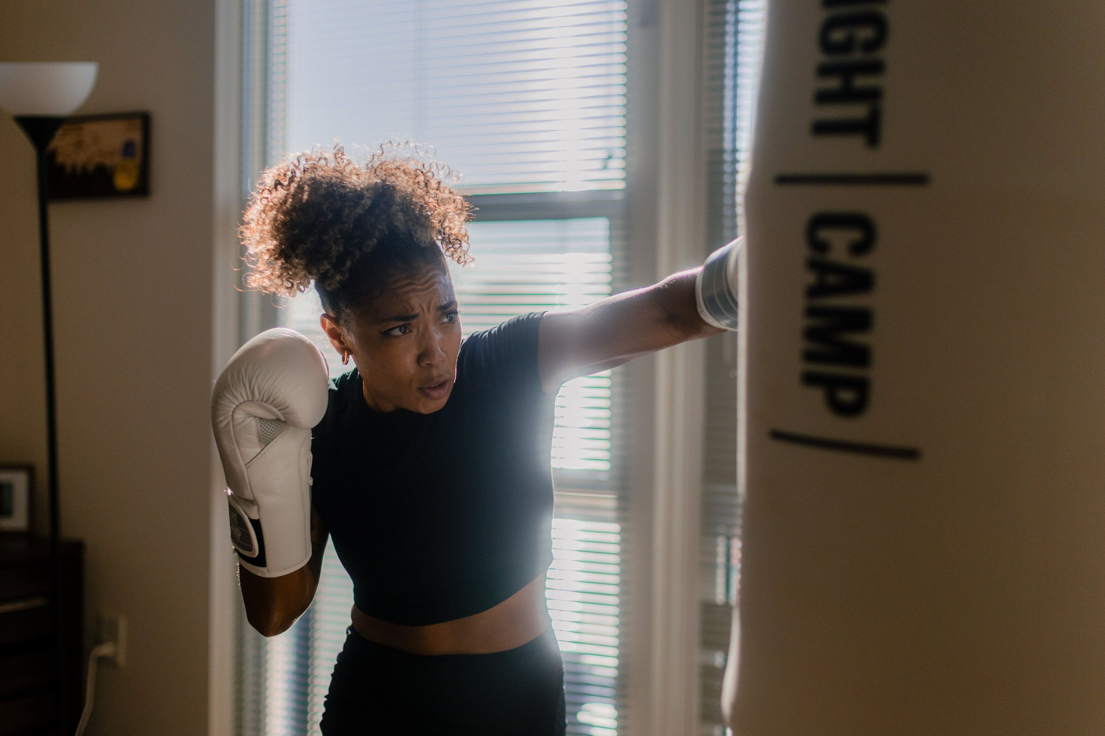Woman boxing indoors, wearing white gloves, focused on punching a FightCamp bag in a sunlit room.