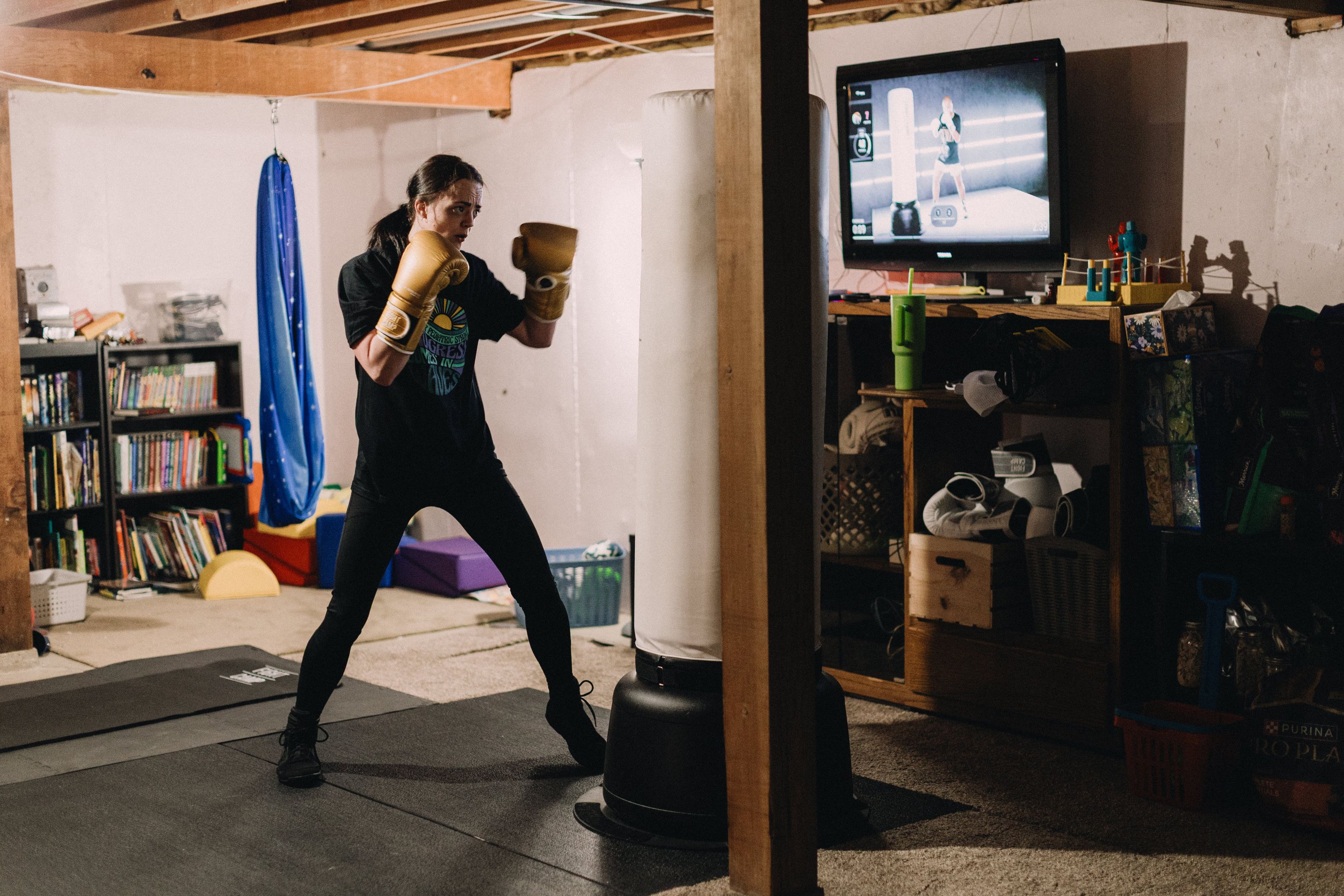 Person wearing boxing gloves practices with a punching bag in a basement, with a TV displaying a workout video in the background.