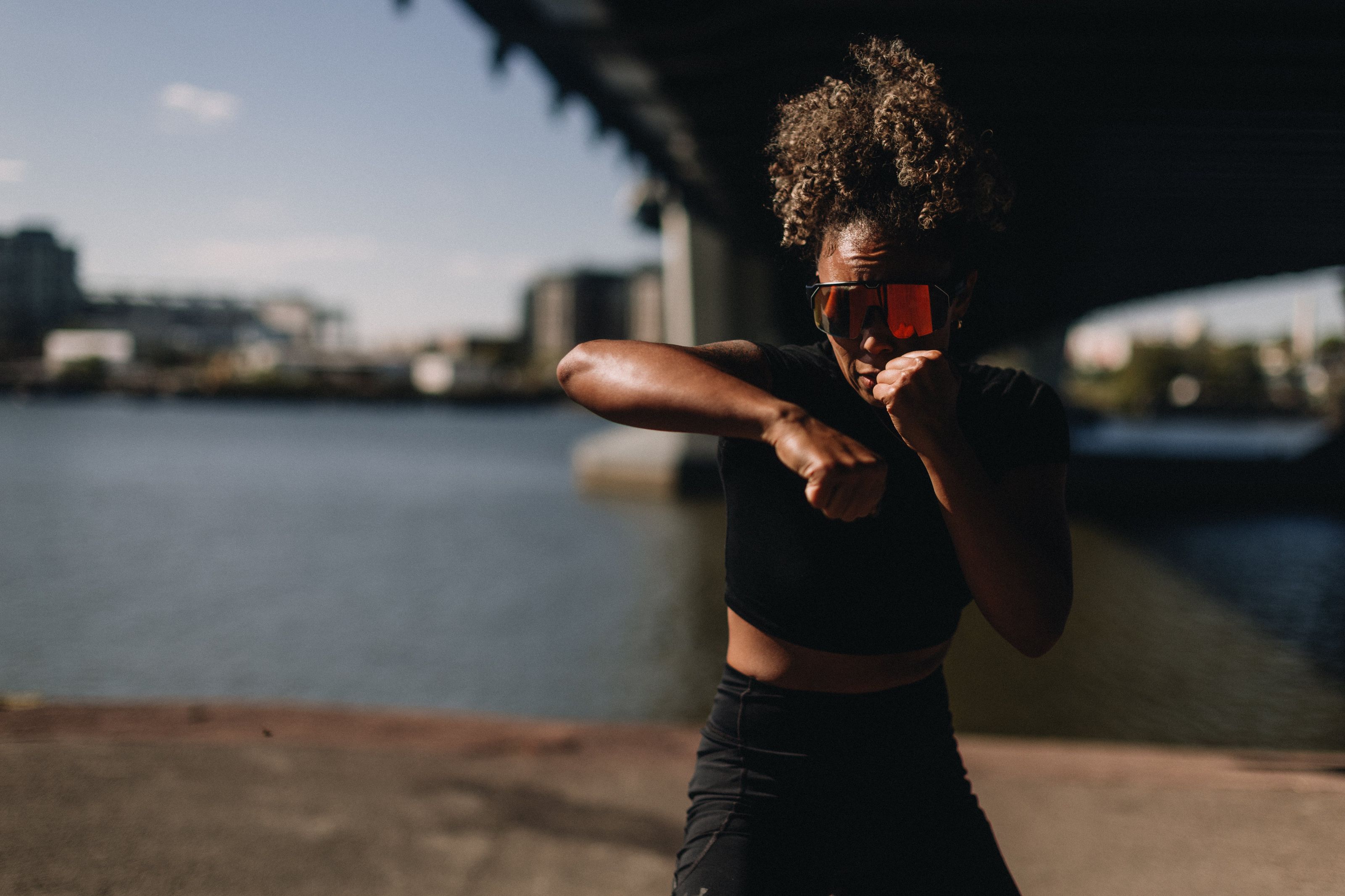 Person shadowboxing near a waterfront under a bridge, wearing a black outfit and reflective sunglasses, with a cityscape in the background.