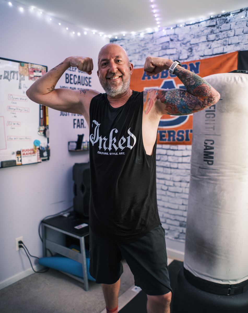 Man in a sleeveless shirt flexing his arms in a room with a FightCamp punching bag, motivational posters, and string lights.