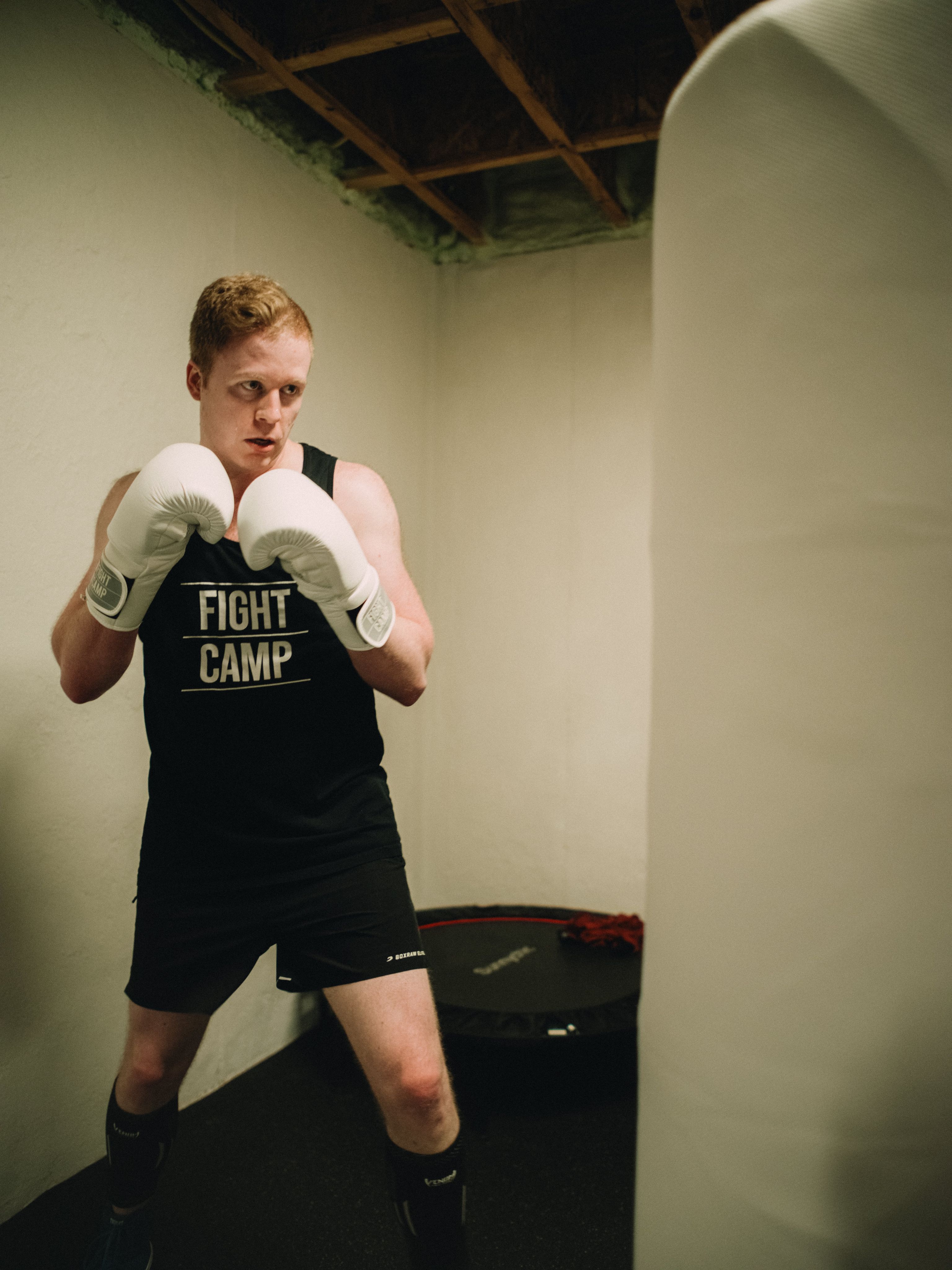 Person in a "Fight Camp" tank top and boxing gloves stands in a fighting stance near a punching bag in a basement setting.