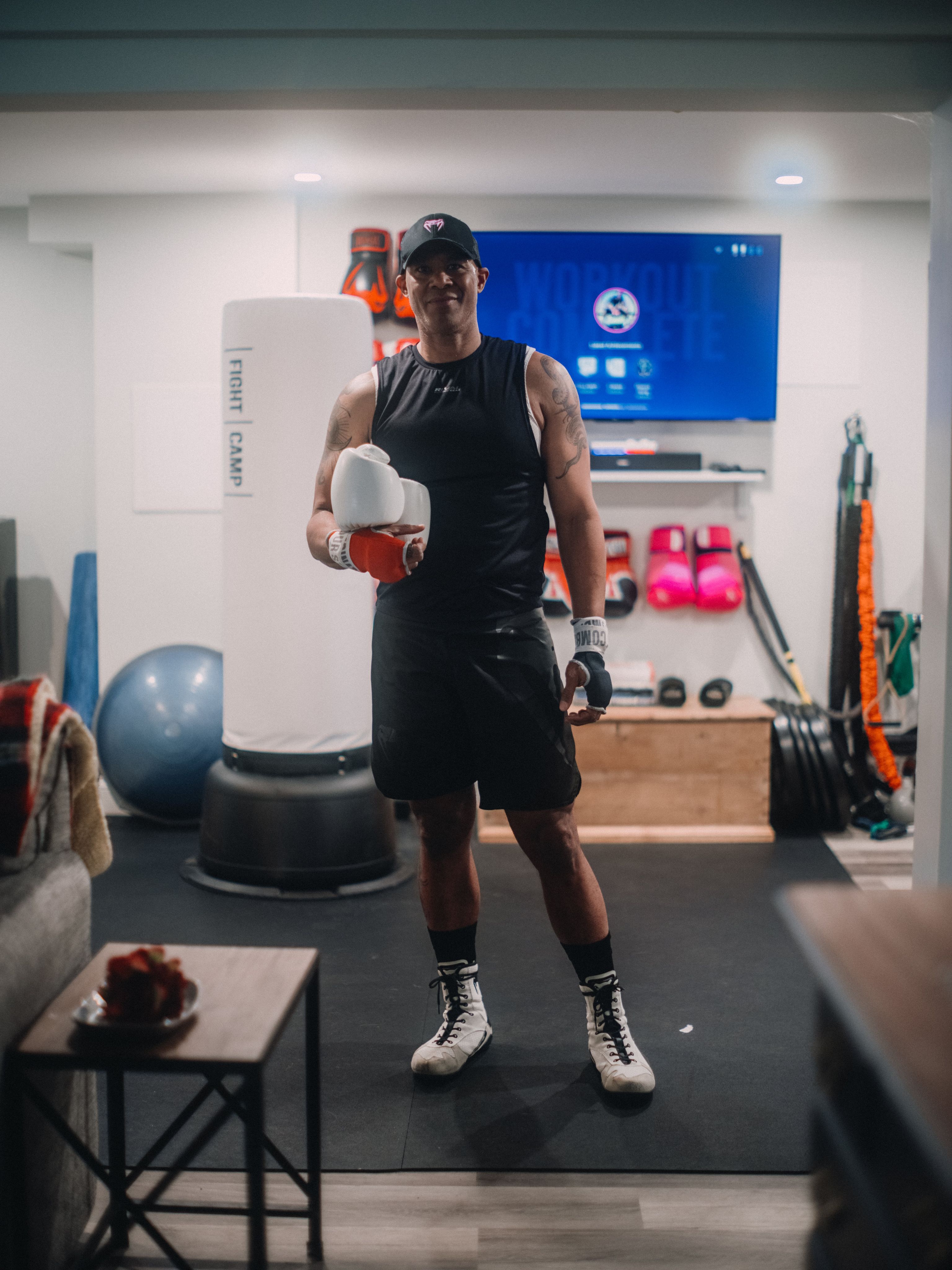 A person in a black tank top and shorts stands in a home gym holding FightCamp boxing gloves, with exercise equipment and a TV in the background.