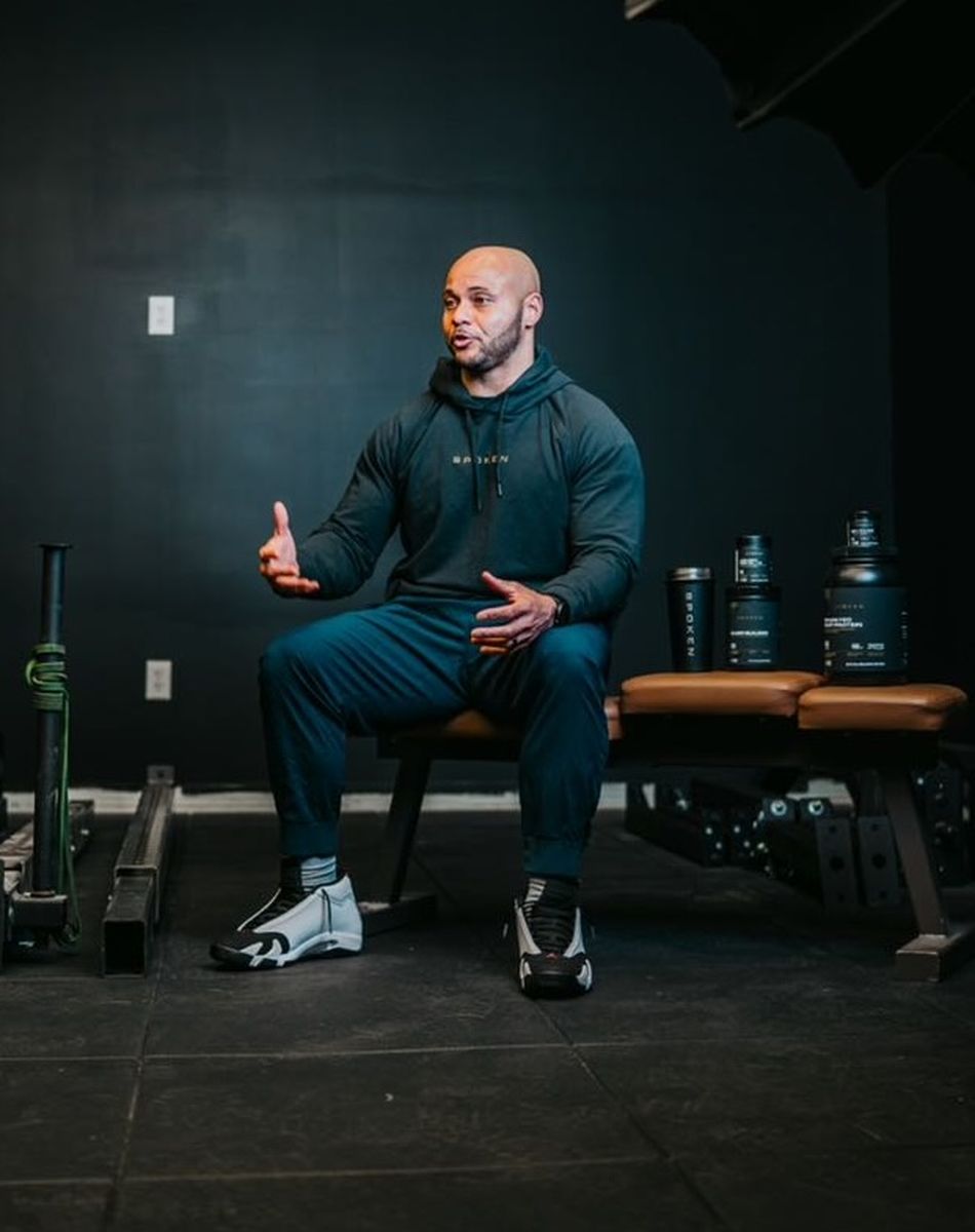 Man in a hoodie and athletic wear gestures while sitting on a bench in a gym with exercise equipment and supplements nearby.