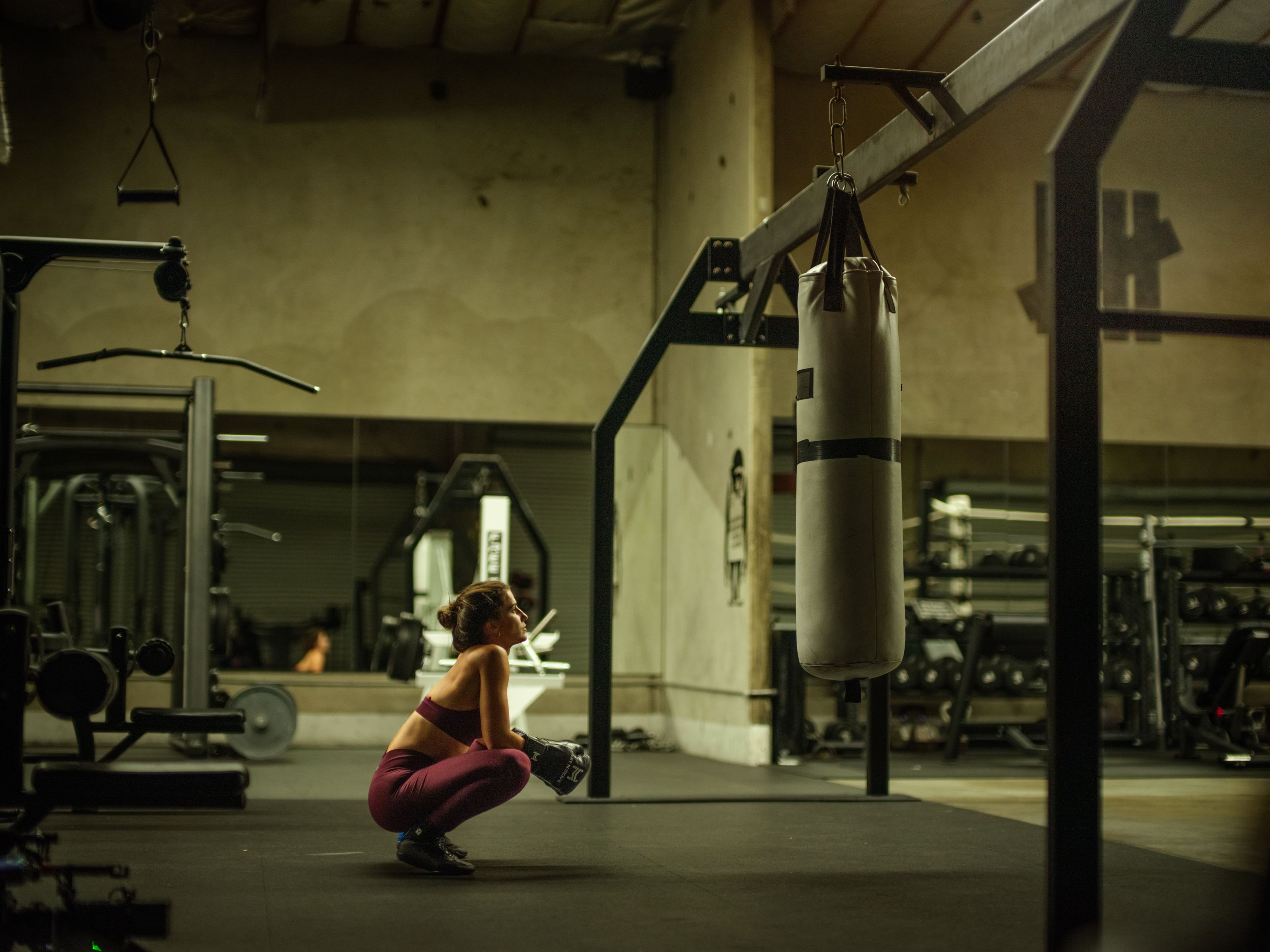 A woman in workout attire squats near a punching bag in a dimly lit gym, surrounded by various exercise equipment.
