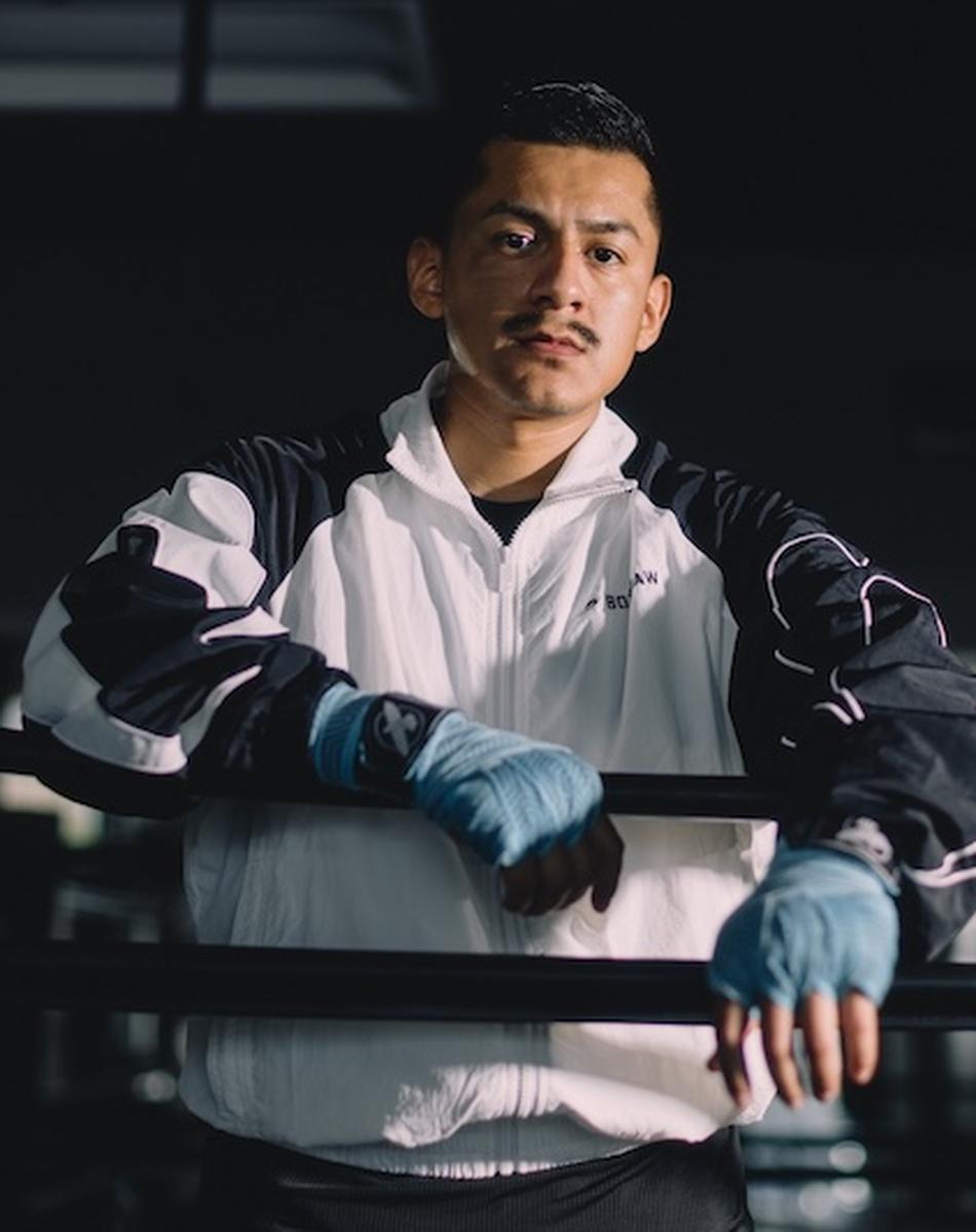 A boxer with wrapped hands leans on the ropes in a dimly lit gym, wearing a black and white jacket, looking confidently at the camera.