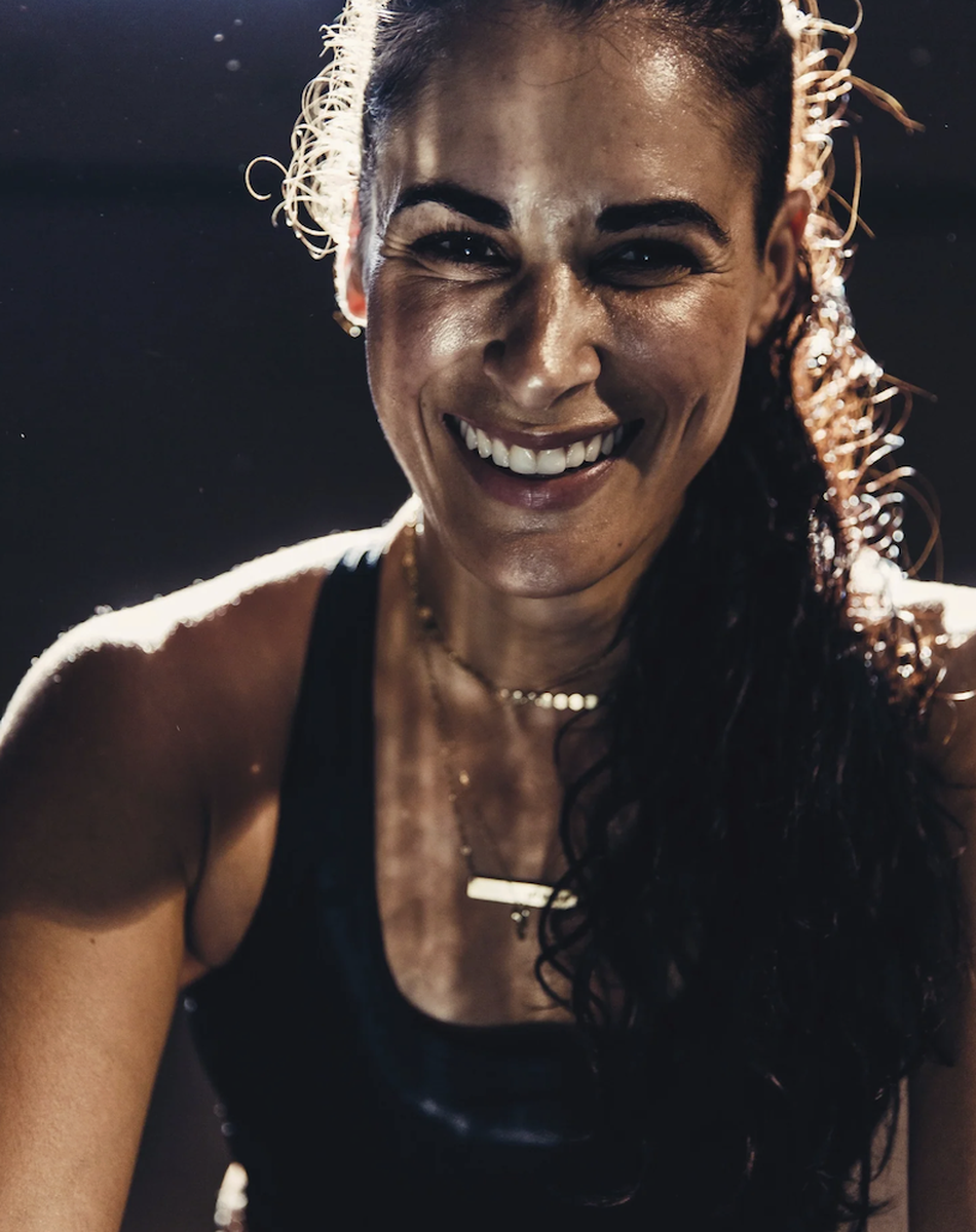 Smiling woman glowing skin in dramatic lighting, wearing a black tank top and necklaces.