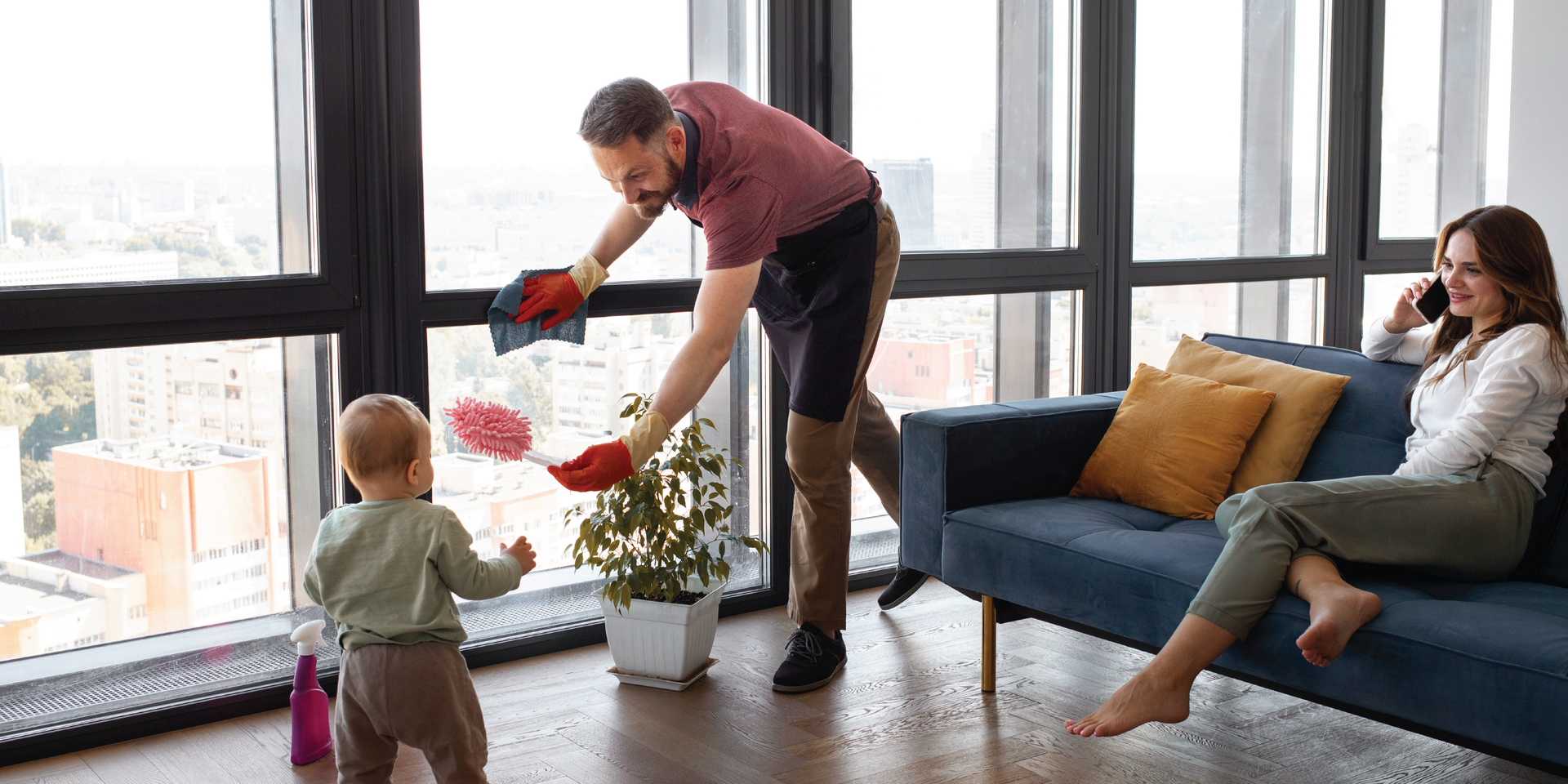 Man with cleaning supplies playing with kid while mom sits on sofa on the phone